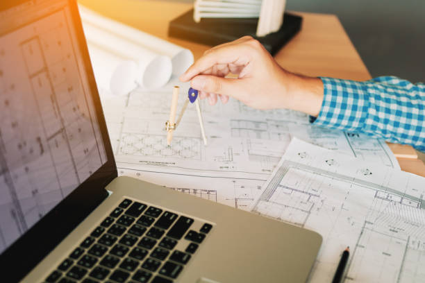 Architect working on blueprint in workplace with laptop and drawing compass.