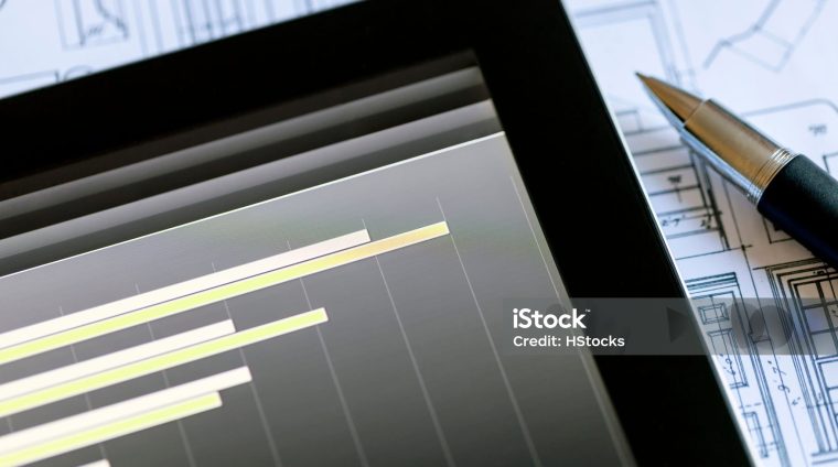 Analyzing stock market from a digital tablet over a detailed diagram with some blue prints and a pen at the background.
